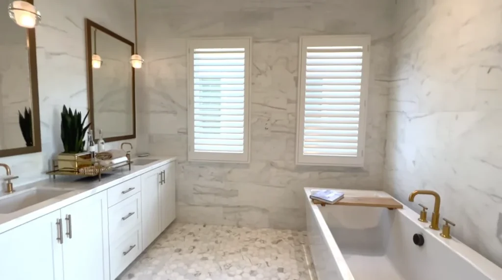 Luxury bathroom with Carrara marble walls, white double vanity, gold fixtures, plantation shutters, and built-in tub